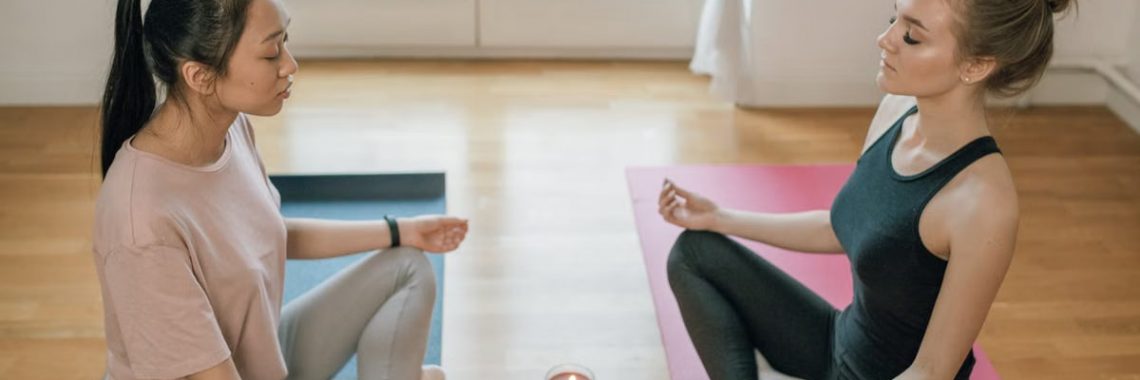 Two women practicing meditation indoors, sitting cross-legged on yoga mats beside lit candles.
